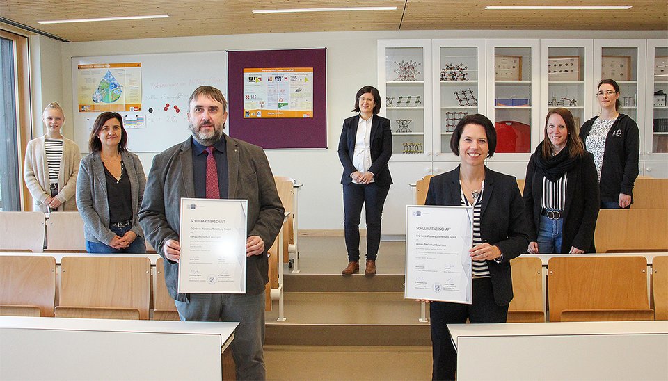 Sechs Personen stehen in einem Klassenzimmer, zwei halten Zertifikate mit dem Titel "Schulpartnerschaft," während im Hintergrund ein Whiteboard und Vitrinen zu sehen sind.