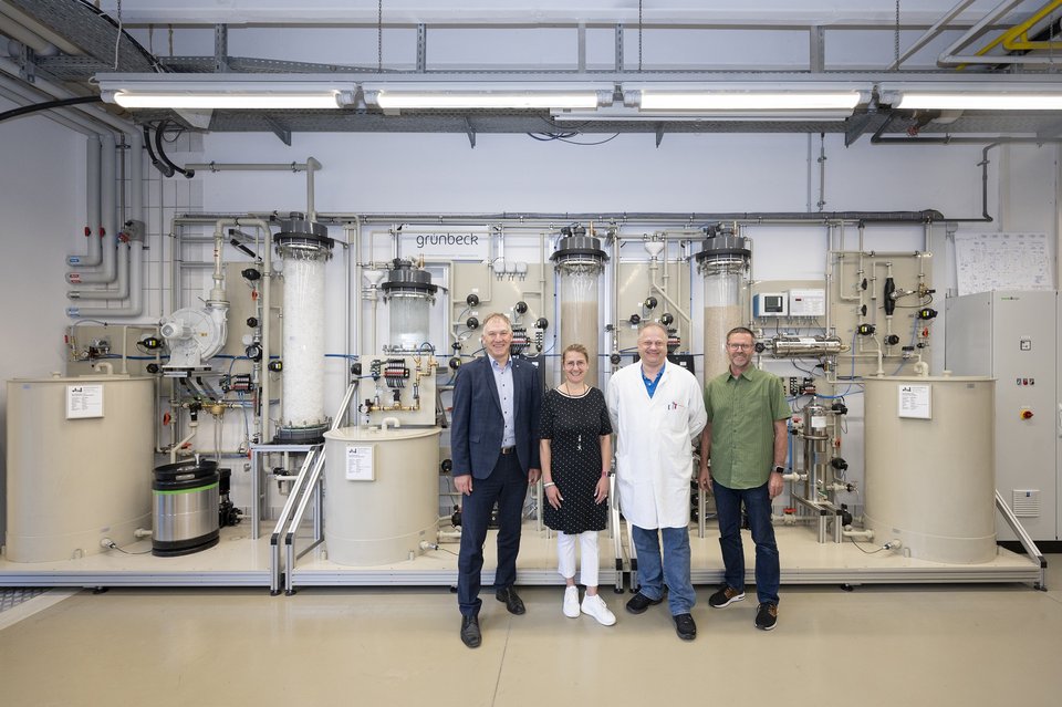 Vier Personen stehen vor einer industriellen Anlage mit verschiedenen Tanks und Rohren; im Hintergrund ist das Logo "grunbeck" zu sehen.
