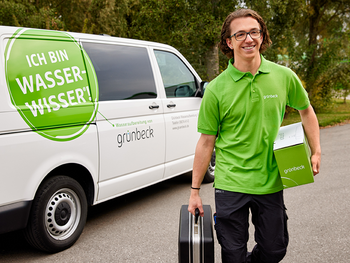 Ein Mann in grüner Arbeitskleidung trägt eine Werkzeugkiste und einen Karton vor einem weißen Lieferwagen mit der Aufschrift "ICH BIN WASSER-WISSER!" und "grünbeck".
