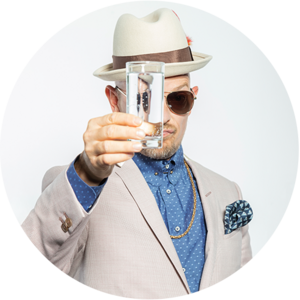 Ein Mann in einem beigen Anzug, Hut und Sonnenbrille hält ein Glas Wasser hoch, im Hintergrund ein neutraler, heller Hintergrund.