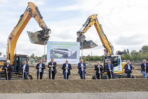 Zehn Personen halten Spaten vor einer Baustelle mit zwei Baggern; ein Schild zeigt "Neubau Wareneingang von Grünbeck" mit einem Gebäudeentwurf.