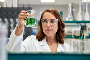 Eine Wissenschaftlerin im Labor hält ein Becherglas mit grüner Flüssigkeit hoch und trägt dabei einen weißen Laborkittel und Schutzbrille.