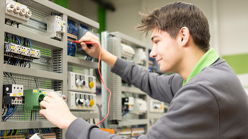 Ein junger Mann arbeitet an einer Schalttafel mit Elektrokabeln und einem Messgerät in einer Werkstattumgebung.