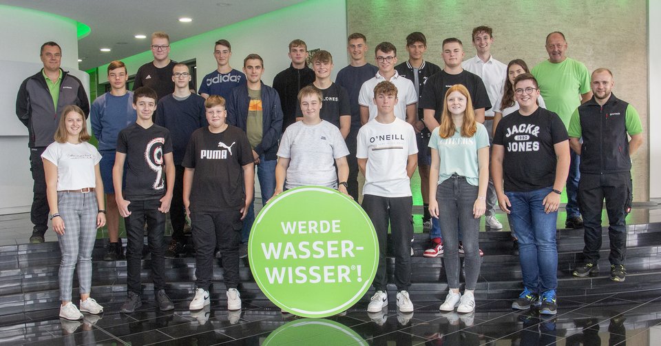 Eine Gruppe junger Menschen steht in einem Gebäude und posiert hinter einem großen grünen Kreis mit der Aufschrift: "WERDE WASSERWISSER!"