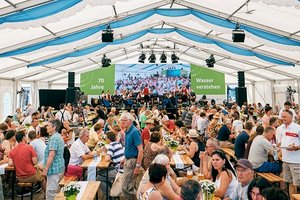 Menschen sitzen und stehen in einem großen Festzelt an Holzbänken und Tischen, während eine Bühne im Hintergrund eine Veranstaltung mit dem Text "70 Jahre Wasser verstehen" zeigt.