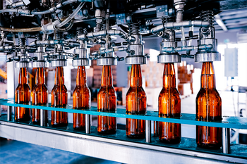 Braune Glasflaschen werden in einer Produktionslinie abgefüllt, umgeben von industriellen Maschinen und Rohren in einer Fabrikumgebung.