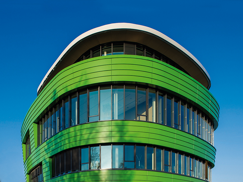 Ein modernes, ovales Gebäude mit grüner Fassade und großen, durchgehenden Fenstern steht vor einem klaren, blauen Himmel.