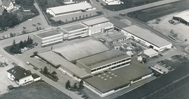 Luftbild einer großen Industrieanlage mit mehreren rechteckigen Gebäuden, umgeben von Straßen und Grünflächen in einer städtischen Umgebung.