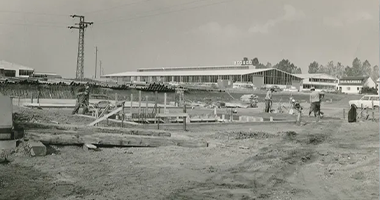 Altes Bild einer Baustelle mit Baumaterialien und arbeitenden Menschen im Vordergrund, im Hintergrund ein großes Gebäude mit moderner Architektur, umgeben von Bäumen und einem Strommast.