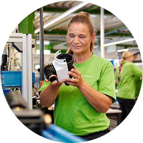 Eine Frau in einem grünen „grünbeck“-Shirt untersucht ein technisches Bauteil in einer industriellen Werkstattumgebung.