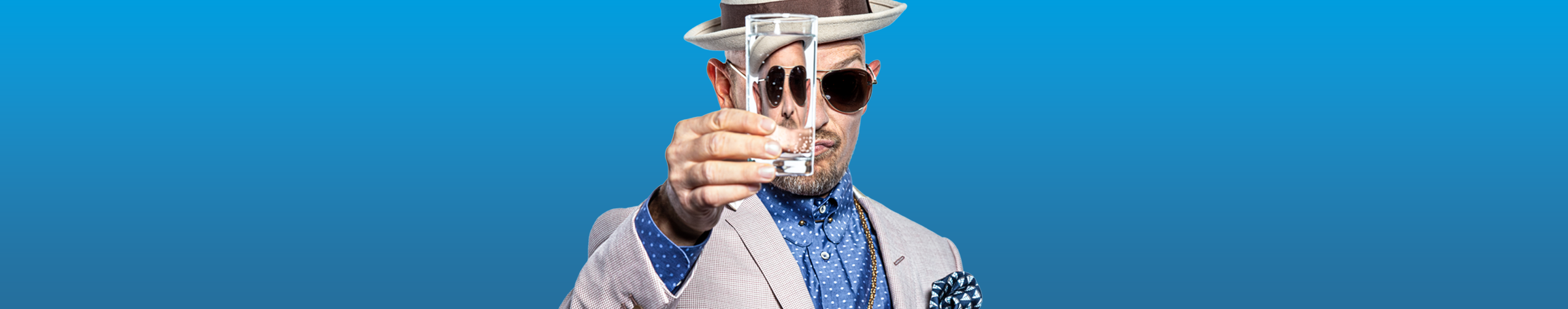 Ein Mann in einem eleganten Anzug und Hut hält ein Glas nach vorne, vor einem einfarbigen blauen Hintergrund.