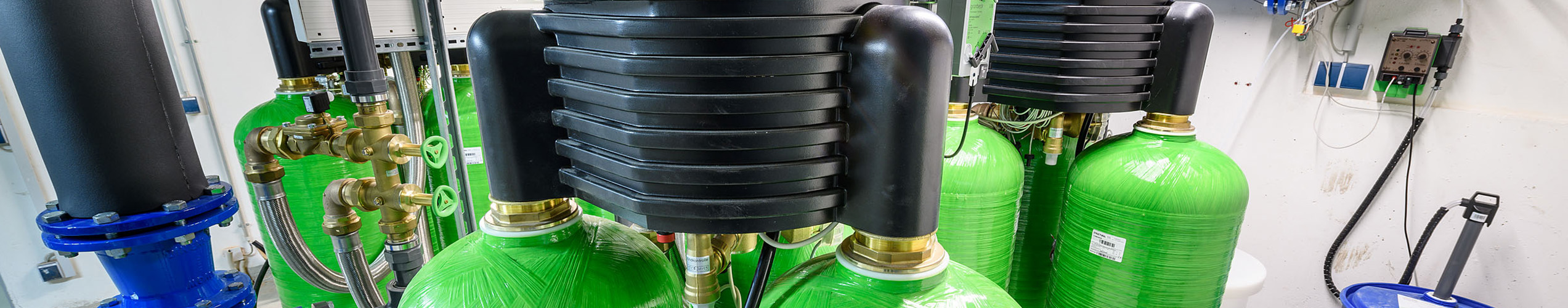 Grüne Druckbehälter mit schwarzen Deckeln sind in einem technischen Raum mit Rohren und Kabeln angeordnet.