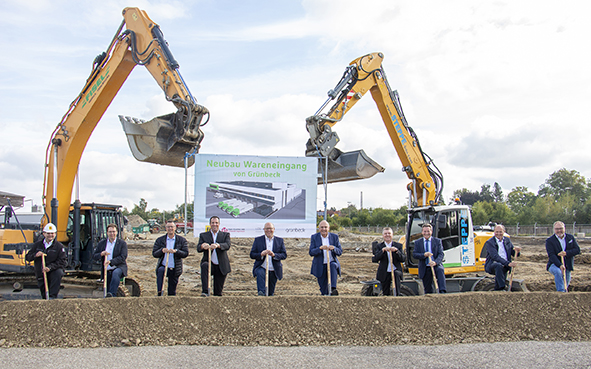 Zehn Personen halten Spaten vor einer Baustelle mit zwei Baggern; ein Schild zeigt "Neubau Wareneingang von Grünbeck" mit einem Gebäudeentwurf.