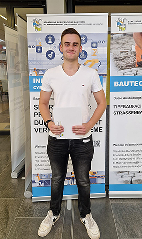 Ein junger Mann hält ein Blatt Papier in den Händen, steht vor zwei Roll-Up-Bannern mit Informationen über Ausbildungsgänge und Bauprojekte in einer Eingangshalle.