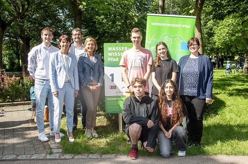 Eine Gruppe von neun Menschen steht lächelnd im Freien vor zwei Schildern mit der Aufschrift "WASSER-WISSER-Pfad" in einem grünen, von Bäumen umgebenen Park.